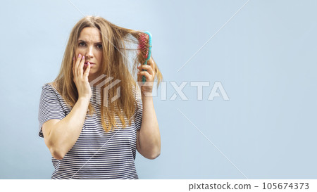 Very sad and upset young woman looking with shock at her damaged hair on blue background Very sad and upset young woman looking with shock at her damaged hair on blue background 105674373
