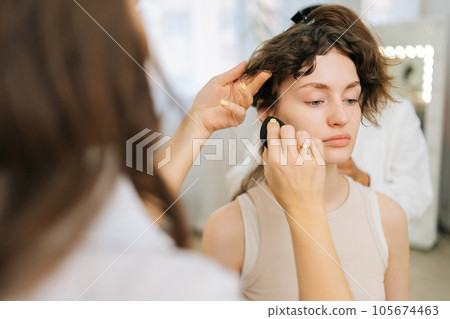 Closeup rear view of unrecognizable female makeup artist applying fluid foundation on young woman face using black sponge. Visagiste applying cosmetic concealer base. Concept of fashion and beauty. Closeup rear view of unrecognizable female makeup artist applying fluid foundation on young woman face using black sponge. Visagiste applying cosmetic concealer base. Concept of fashion and beauty. 105674463