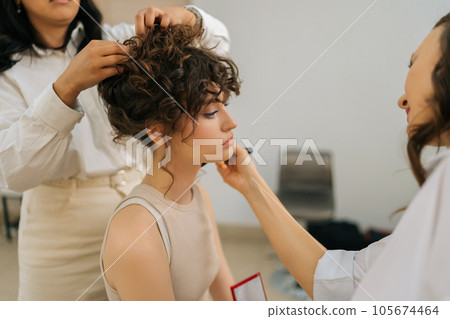 Close-up side view of makeup artist applying toner on face of beautiful model. Hairdresser making hair style in tandem, giving beauty salon service and treatment. Concept of fashion and beauty. Close-up side view of makeup artist applying toner on face of beautiful model. Hairdresser making hair style in tandem, giving beauty salon service and treatment. Concept of fashion and beauty. 105674464