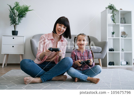 Tween girl and mom holding gaming consoles in room interior Tween girl and mom holding gaming consoles in room interior 105675796