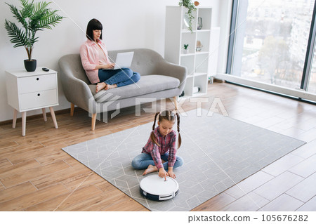 Female parent working while kid turning on robot vacuum 105676282