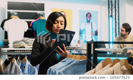Store manager working on boutique stock with tablet, analyzing all clothes on hangers and racks. Female employee doing inventory in clothing store, examining merchandise. Handheld shot. Store manager working on boutique stock with tablet, analyzing all clothes on hangers and racks. Female employee doing inventory in clothing store, examining merchandise. Handheld shot. 105677625