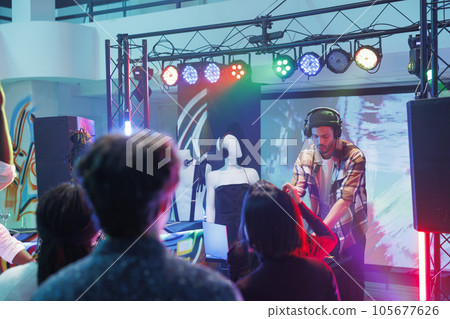People attending techno music concert and enjoying nightlife in club illuminated with lights. Man dj playing and mixing tracks for crowd at discotheque party in nightclub People attending techno music concert and enjoying nightlife in club illuminated with lights. Man dj playing and mixing tracks for crowd at discotheque party in nightclub 105677626
