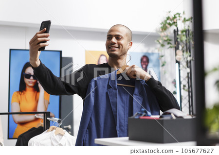 Smiling arab man holding hanger with stylish jacket while taking selfie using smartphone front camera in clothing store. Blogger showcasing apparel to social media channel followers in boutique Smiling arab man holding hanger with stylish jacket while taking selfie using smartphone front camera in clothing store. Blogger showcasing apparel to social media channel followers in boutique 105677627