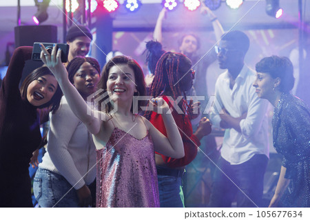 Smiling woman taking selfie while dancing with friends at nightclub discotheque. Happy girl holding mobile phone and posing for photo on club dancefloor illuminated with stage spotlights Smiling woman taking selfie while dancing with friends at nightclub discotheque. Happy girl holding mobile phone and posing for photo on club dancefloor illuminated with stage spotlights 105677634