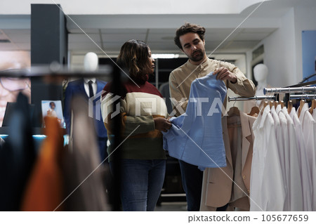 Clothing store customer getting fashion recommendation from consultant while browsing trendy apparel rack. Woman and boutique worker holding shirt on hanger and selecting size Clothing store customer getting fashion recommendation from consultant while browsing trendy apparel rack. Woman and boutique worker holding shirt on hanger and selecting size 105677659
