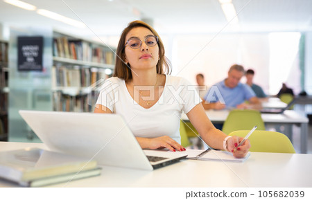Confident student girl studying in the university library on a laptop, writes an important synopsis in copybook Confident student girl studying in the university library on a laptop, writes an important synopsis in copybook 105682039