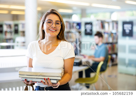 Joyful young Latin female reading the book while standing in library 105682159