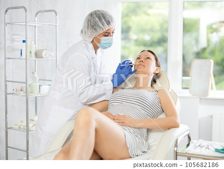 Doctor in disposable mask performs procedure of lip augmentation of girl patient in clinic. 105682186