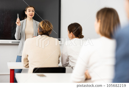 Young female business coach explaining topic while standing against group of people sitting at desks in front of her 105682219