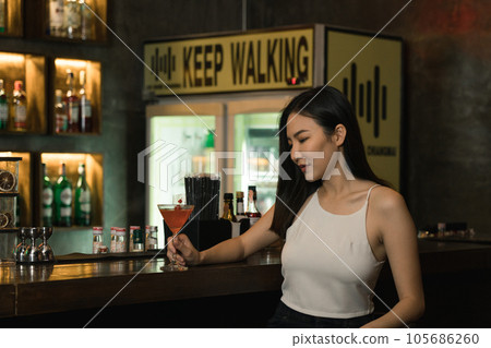 Asian woman drinking a cocktail at a bar at night. Asian woman drinking a cocktail at a bar at night. 105686260