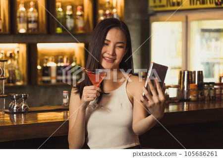 Asian woman taking a photo of herself while drinking whiskey at the bar. 105686261