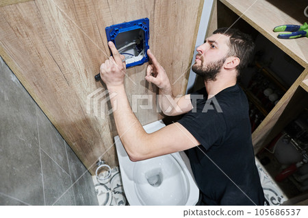 Bearded man holding screwdriver tool while installing toilet flush panel in apartment under renovation. Male plumber adjusting frame of toilet actuator panel mounted on the wall. Bearded man holding screwdriver tool while installing toilet flush panel in apartment under renovation. Male plumber adjusting frame of toilet actuator panel mounted on the wall. 105686537