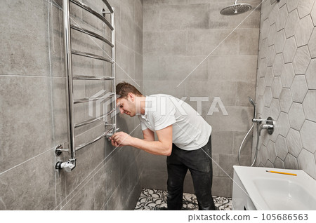 Man hanging electric towel rack dryer on the wall with ceramic tile in bathroom. Male worker installing bath towel heater in apartment under renovation. Concept of home renovation. Man hanging electric towel rack dryer on the wall with ceramic tile in bathroom. Male worker installing bath towel heater in apartment under renovation. Concept of home renovation. 105686563