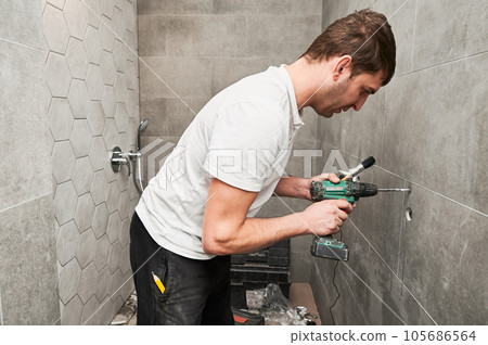 Male worker using electric drilling machine and construction brush while working on bathroom renovation. Male plumber drilling wall with power drill while installing shower system at home. Male worker using electric drilling machine and construction brush while working on bathroom renovation. Male plumber drilling wall with power drill while installing shower system at home. 105686564