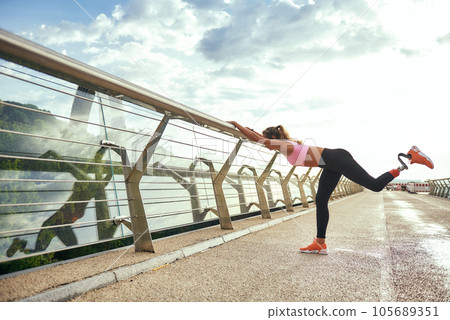 Morning workout. Disabled athletic woman with prosthetic leg in sportswear doing sport exercises while standing on the bridge. Side view 105689351