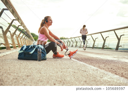 Time to relax. Side view of young beautiful woman in headphones with leg prosthesis listening music and looking aside with smile while sitting on the bridge Time to relax. Side view of young beautiful woman in headphones with leg prosthesis listening music and looking aside with smile while sitting on the bridge 105689397