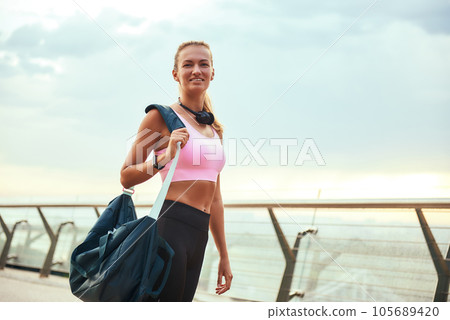 Full of energy. Beautiful young woman in sports clothing carrying her sport bag and smiling while standing on the bridge in the morning Full of energy. Beautiful young woman in sports clothing carrying her sport bag and smiling while standing on the bridge in the morning 105689420