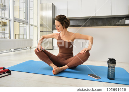 Sport and wellbeing. Young brunette fitness girl, wearing activewear, sitting on yoga mat, meditating, practice relaxation and mindfulness at home, workout in kitchen 105691656