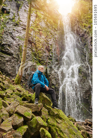 Tourist attraction of Germany - falls of Burgbach Waterfall near Schapbach, Black Forest, Baden-Wurttemberg, Germany. Man hiker in blue jacket standing on stone and looks at flow of falling water Tourist attraction of Germany - falls of Burgbach Waterfall near Schapbach, Black Forest, Baden-Wurttemberg, Germany. Man hiker in blue jacket standing on stone and looks at flow of falling water 105691695