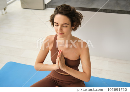 Young healthy beautiful woman in sports top and leggings practicing yoga at home, sitting in lotus position on yoga mat, meditating, smiling, relaxed, eyes closed, meditation concept Young healthy beautiful woman in sports top and leggings practicing yoga at home, sitting in lotus position on yoga mat, meditating, smiling, relaxed, eyes closed, meditation concept 105691730