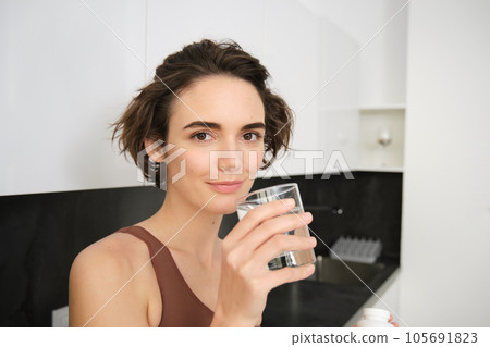 Healthy lifestyle and female wellbeing. Young beautiful woman standing in kitchen and drinking glass of water after workout training. Fitness girl stays hydrated after gym class 105691823