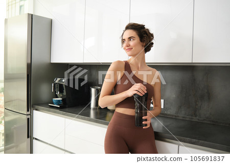 Sport and healthy lifestyle. Portrait of smiling fitness woman in activewear, standing near kitchen counter at home, drinking from water bottle, preparing for workout gym training Sport and healthy lifestyle. Portrait of smiling fitness woman in activewear, standing near kitchen counter at home, drinking from water bottle, preparing for workout gym training 105691837