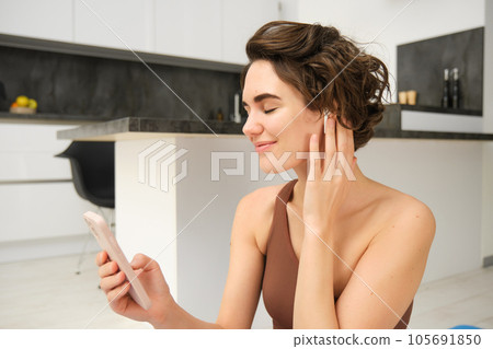 Portrait of beautiful, smiling fitness woman, looks at smartphone, video calls fitness instructor, online gym classes from home, looks at phone and wears wireless headphones, workout remotely Portrait of beautiful, smiling fitness woman, looks at smartphone, video calls fitness instructor, online gym classes from home, looks at phone and wears wireless headphones, workout remotely 105691850
