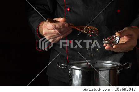 The cook adds fragrant dry peppers to a pot of boiling food. Restaurant kitchen cooking concept with advertising space on black background. Spoon in the chef hand. 105692013