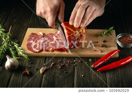 Close-up of a cook's hands with a knife cuts smoked veal meat into thin slices for a sandwich. Fast food concept on dark background. Close-up of a cook's hands with a knife cuts smoked veal meat into thin slices for a sandwich. Fast food concept on dark background. 105692029