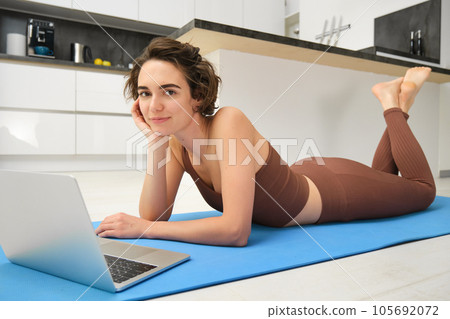 Portrait of beautiful sportswoman, fitness girl watching videos on laptop during workout, follow online pilates videos while exercise at home, wearing activewear in kitchen Portrait of beautiful sportswoman, fitness girl watching videos on laptop during workout, follow online pilates videos while exercise at home, wearing activewear in kitchen 105692072