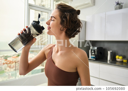 Healthy lifestyle and sport. Smiling brunette female athlete, fitness girl drinks water from gym bottle with pleased face, standing at home in kitchen Healthy lifestyle and sport. Smiling brunette female athlete, fitness girl drinks water from gym bottle with pleased face, standing at home in kitchen 105692080