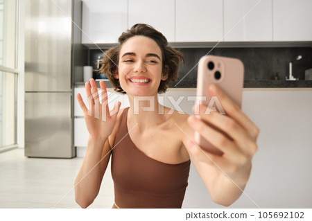 Beautiful young woman using mobile phone and workout at home, waving hand at smartphone, video chats, doing yoga training live online, records sports vlog in her house Beautiful young woman using mobile phone and workout at home, waving hand at smartphone, video chats, doing yoga training live online, records sports vlog in her house 105692102