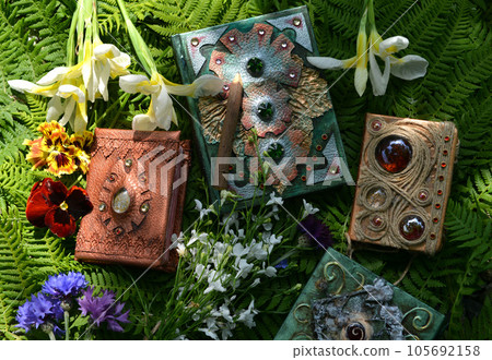 Still life with beautiful magic book of spells or diary with flowers against green leaves. Occult, esoteric and witchcraft concept. Mystic wicca background with vintage objects Still life with beautiful magic book of spells or diary with flowers against green leaves. Occult, esoteric and witchcraft concept. Mystic wicca background with vintage objects 105692158