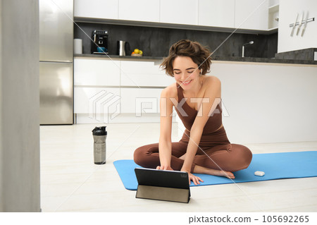 Young fit and healthy woman, yoga instructor, fitness girl in tracksuit, sits on floor rubber mat, using digital tablet, watches video pilates tutorial, stretching exercises 105692265