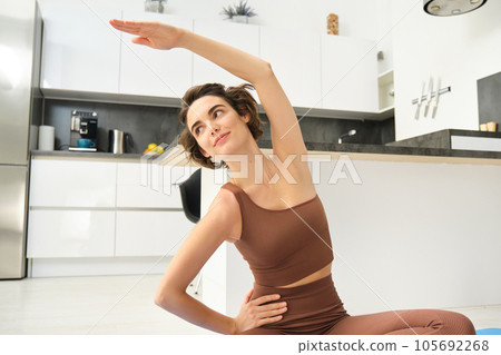 Fitness girl doing exercises at home. Young woman athlete sitting on yoga mat in bright room, stretching her arms, doing pilates exercises 105692268