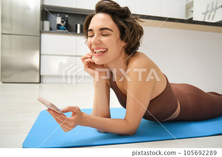 Close up portrait of fitness girl, lying on yoga mat and laughing, using smartphone, records workout exercises on mobile phone, does training at home Close up portrait of fitness girl, lying on yoga mat and laughing, using smartphone, records workout exercises on mobile phone, does training at home 105692289