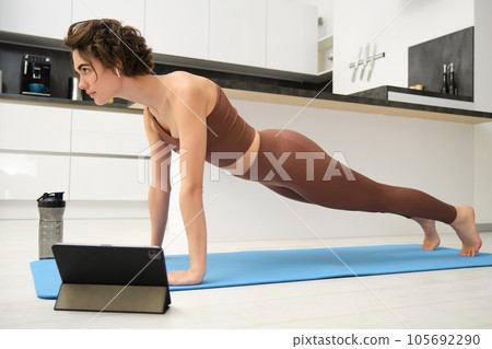 Workout, training and sports concept. Young serious woman follows online training video, stands in plank on rubber yoga mat at home, boosts endurance, excerice on abs 105692290