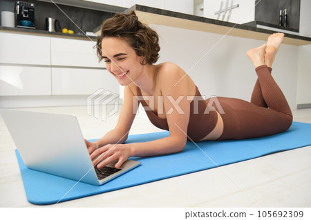 Portrait of woman athlete, girl workout at home, watches fitness videos on laptop, lying on rubber mat and doing indoor yoga session Portrait of woman athlete, girl workout at home, watches fitness videos on laptop, lying on rubber mat and doing indoor yoga session 105692309