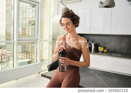 Sport and healthy lifestyle. Portrait of smiling fitness woman in activewear, standing near kitchen counter at home, drinking from water bottle, preparing for workout gym training 105692310
