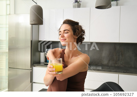 Happy brunette sportswoman, girl in activewear drinking glass of orange juice, sitting in her kitchen counter and smiling Happy brunette sportswoman, girl in activewear drinking glass of orange juice, sitting in her kitchen counter and smiling 105692327