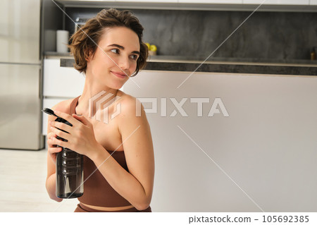 Fitness instructor, smiling young woman workout at home, sits on yoga mat and drinks water, does exercises, training session in her kitchen 105692385