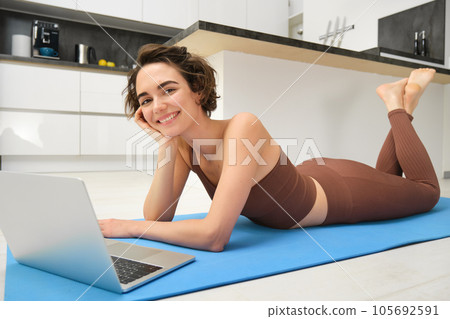 Portrait of beautiful sportswoman, fitness girl watching videos on laptop during workout, follow online pilates videos while exercise at home, wearing activewear in kitchen 105692591