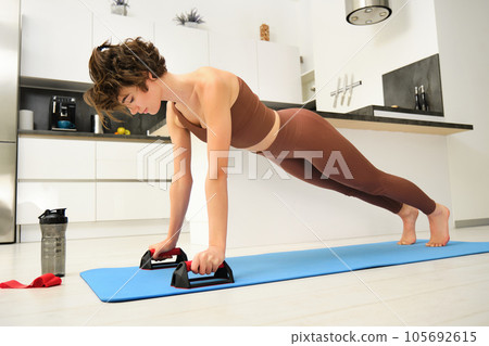 Young fitness woman, stands on push up bars stand, doing plunk exercise at home, does chest press workout training in her kitchen, exercising on yoga rubber mat Young fitness woman, stands on push up bars stand, doing plunk exercise at home, does chest press workout training in her kitchen, exercising on yoga rubber mat 105692615