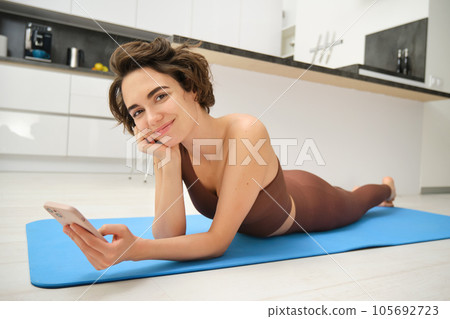 Cute brunette woman lying on yoga mat at home, doing fitness training alone indoors, holding smartphone and smiling at camera 105692723