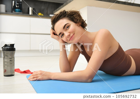 Sport and healthy lifestyle. Portrait of brunette sportswoman doing fitness in activewear, lying on rubber mat on floor, doing workout training in kitchen, drinking water from gym bottle Sport and healthy lifestyle. Portrait of brunette sportswoman doing fitness in activewear, lying on rubber mat on floor, doing workout training in kitchen, drinking water from gym bottle 105692732