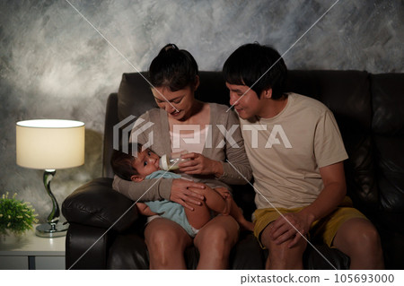 parents (father and mother) feeding milk bottle to infant baby on sofa in the living room at night parents (father and mother) feeding milk bottle to infant baby on sofa in the living room at night 105693000