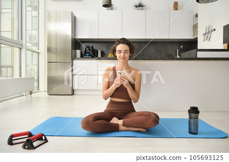 Yoga and sport at home concept. Young woman sitting in bright rome with mobile phone, doing workout on rubber mat, searching for exercises online on smartphone app, using sports equipment Yoga and sport at home concept. Young woman sitting in bright rome with mobile phone, doing workout on rubber mat, searching for exercises online on smartphone app, using sports equipment 105693125