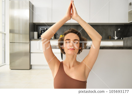 Image of young woman meditating, raising her hands up above head, practice mindfulness at home, doing yoga session on rubber mat in bright room, wearing sportswear Image of young woman meditating, raising her hands up above head, practice mindfulness at home, doing yoga session on rubber mat in bright room, wearing sportswear 105693177