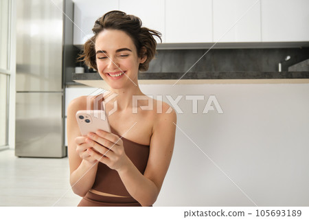 Sport and women lifestyle. Young woman does workout at home, sits on yoga mat with smartphone in bright kitchen, wears activewear, uses mobile phone app for indoor exercises 105693189